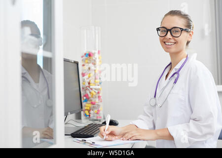 Portrait of smiling female doctor writing on clipboard while sitting at desk in office, copy space Stock Photo