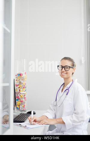 Vertical portrait of smiling female doctor writing on clipboard while sitting at desk in office, white wall copy space Stock Photo