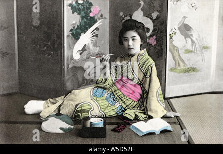 [ 1900s Japan - Japanese Woman Smoking a Pipe ] —   Woman in kimono holding a kiseru pipe. She is sitting on tatami rice mats on the floor in front of a screen.  20th century vintage postcard. Stock Photo