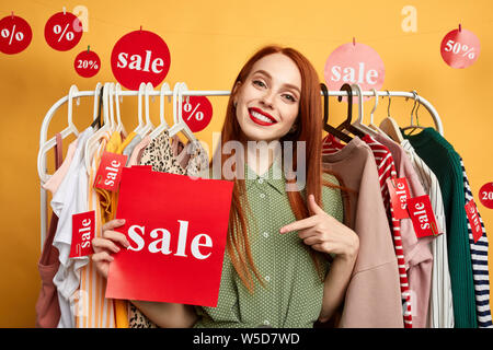 awesome lovely ginger girl in stylish green dress pointing to the sale. close up photo. isolated yellow background. studio shot.hurry up to the shop. Stock Photo