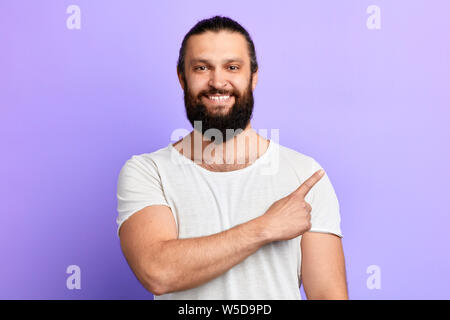 positive bearded guy pointing aside with finger .place for text, copy space. isolated blue background. studio shot Stock Photo
