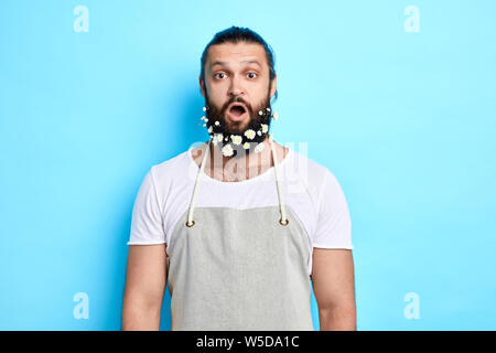 Shocked young man in gray apron with wide open mouth looking at the camera. close up portrait. isolated blue background. studio shot Stock Photo