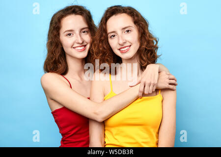 Pleased awesome twins wearing t-shirts embracing each other and looking at the camera over blue background. love, support, friendship Stock Photo
