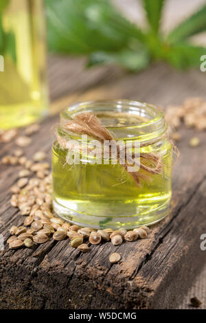 Hemp oil in glass jar and grains of cannabis on old wooden board, bottle with oil and leaves are blurred. Stock Photo