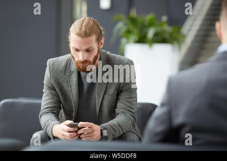 Serious young bearded man in gray jacket sitting in lobby and using internet app on smartphone Stock Photo