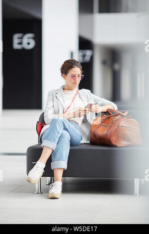 Serious hipster girl in casual outfit sitting on leather sofa with travel bag in airport waiting area and surfing net on phone