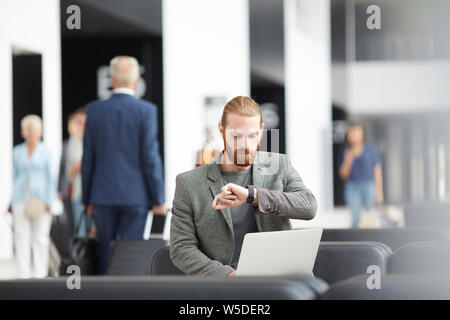 Serious young bearded entrepreneur sitting with laptop in airport and looking at wristwatch while checking time till boarding Stock Photo
