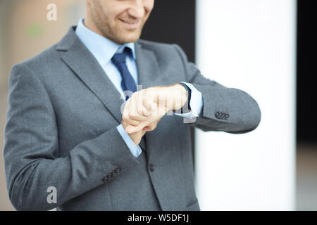 Close-up of smiling businessman in gray suit standing in lobby and checking time on wristwatch Stock Photo
