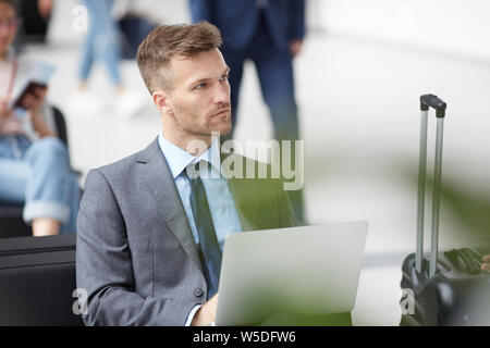 Thoughtful handsome young businessman with stubble sitting with luggage in airport and working with data on laptop Stock Photo