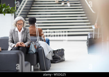 Content elderly Asian lady with gray hair sitting on black leather sofa in airport and browsing internet on smartphone Stock Photo