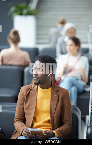 Serious pensive young Afro-American man in brown jacket sitting on sofa in airport and listening to audio in wireless earphones waiting for boarding Stock Photo