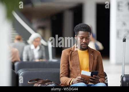 Serious pensive young black man with beard sitting in airport and communicating via online app on phone Stock Photo