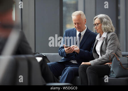 Serious handsome mature man in suit checking notes on smartphone and showing it to Asian colleague in airport Stock Photo