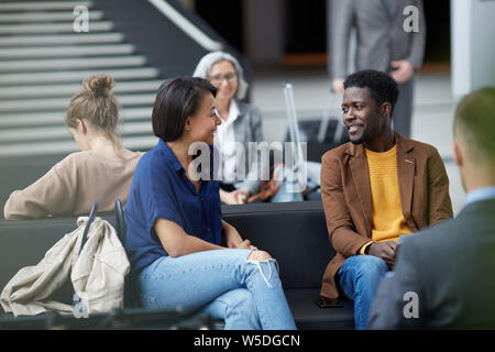 Positive handsome young Afro-American man with beard sitting on sofa and talking to lady in airport area Stock Photo