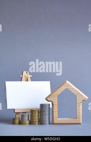 A wooden figure of house with several columns of coins nearby and white board on miniature easel on gray background, the concept of buying or renting Stock Photo