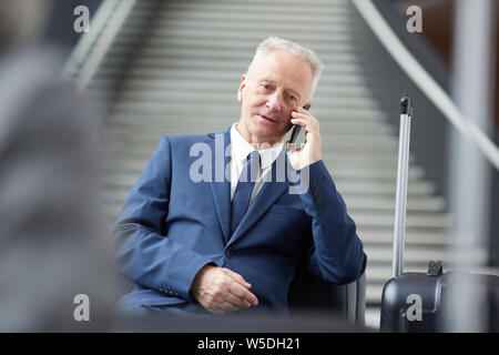 Serious handsome mature businessman in formal suit sitting on sofa in airport and communicating on cellphone Stock Photo