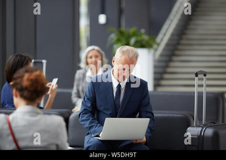 Content handsome gray-haired businessman in formal suit sitting on sofa and examining data on laptop while waiting for boarding Stock Photo