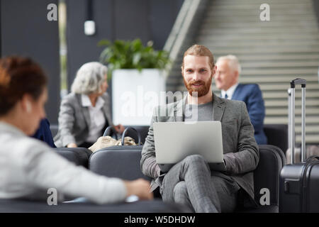 Portrait of smiling red-bearded entrepreneur in gray jacket sitting on sofa and using laptop in airport Stock Photo