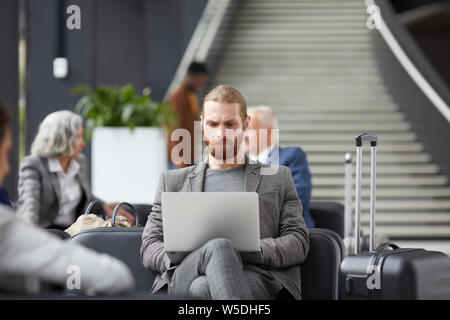 Concentrated bearded guy sitting on sofa and using modern laptop while working in airport Stock Photo