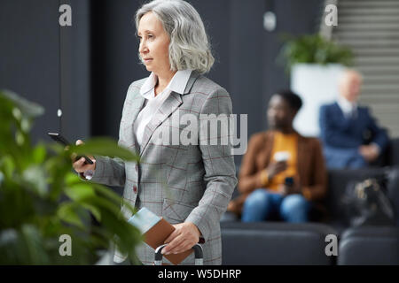 Pensive Asian gray-haired lady in checkered jacket standing with suitcase in waiting area and holding passport while checking information on phone Stock Photo