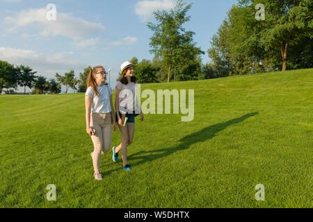 Two teen girls students with backpacks and books walking on green grass in the park, sunny day Stock Photo