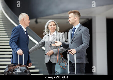 Group of business professionals in formalwear standing in airport and holding suitcases while talking to each other Stock Photo