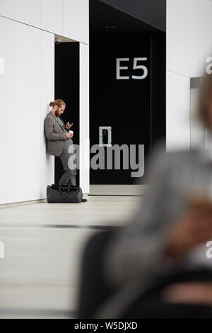 Serious handsome young entrepreneur in jacket standing with journey bag and wasting time in airport Stock Photo
