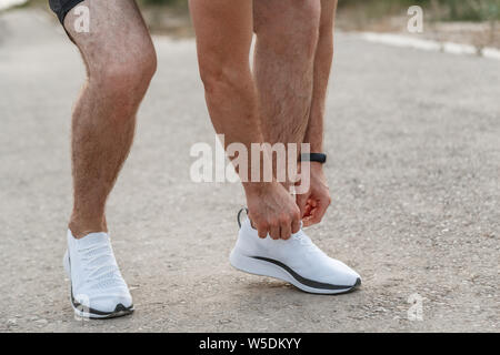 portrait of Caucasian guy in a blue t-shirt and black shorts,who ties his laces on white sports shoes before Jogging Stock Photo