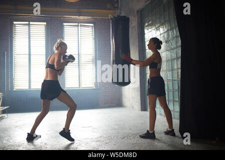 Dramatic backlit portrait of two female boxers hitting punching bag in shabby sports club, copy space Stock Photo
