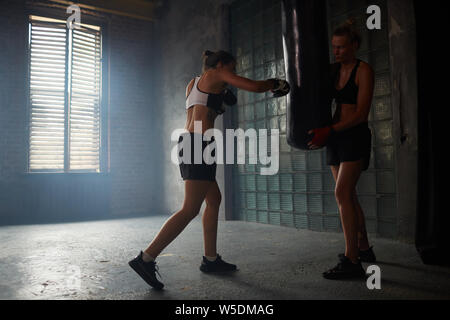Dramatic portrait of forceful young woman hitting punching bag during boxing practice in shabby sports club, copy space Stock Photo