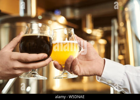 Close up of two male hands holding glasses with beer. Assortment of drinks. Dark and light beer. Industry of brewing beer. Equipment of modern mini brewery on background. Stock Photo