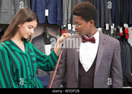 Beautiful female shop consultant choosing suit for young man with curly hair. Brunette holding measuring tape and measuring sleeve of grey jacket on client. Fashion and style. Stock Photo