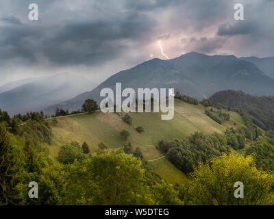 Grim landscape with remote church on hilltop and thunderstorm approaching across mountains in background Stock Photo