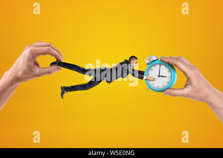 Small businessman being pulled in opposite directions by two big hands, and clutching with his both hands at alarm clock in one of the hands. Stock Photo