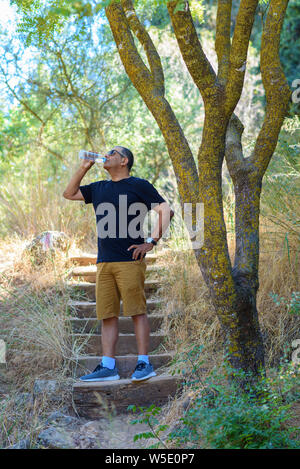 Elderly success athlete senior man drinking water after trip or travel on sunny summer day in forest, antique park. Outdoor portrait mature man tourist,lifestyle, sport, health, retirement concept. Stock Photo