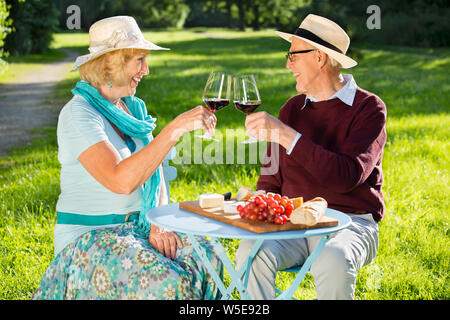 Happy senior couple clinking glasses in the park, looking into each other's eyes flirting, drinking wine, eating cheese with grapes and baguette. Stock Photo