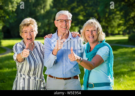 Three senior friends showing optimism, standing arm in arm, smiling enthusiasticallly with thumbs turned up. Stock Photo