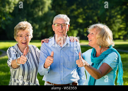 Active senior friends, two women and a man standing arm in arm, smiling and showing the thumbs up, blonde beautiful woman is looking at the handsome o Stock Photo