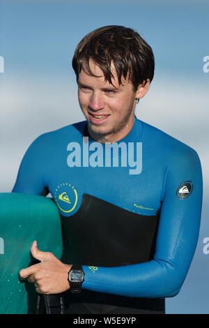 Young man wearing a black and blue wetsuit carrying a blue surfboard in Huntington Beach, California on October 19, 2018 Stock Photo