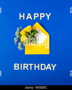 Yellow roses and white Gypsophila in envelope close-up on blue background. Text Happy Birthday, wooden letters. Top view, flat lay. Template for birth Stock Photo