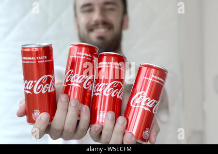 KHARKIV, UKRAINE - JULY 15, 2019: Smiling man holding many non-alcoholic Coca-Cola aluminium tin cans in garage interior. Coca Cola is the most famous Stock Photo