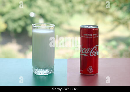 KHARKIV, UKRAINE - JULY 20, 2019: Coca cola red tin can and cup of fresh milk on colored surface with background of green garden. Healthy and unhealth Stock Photo