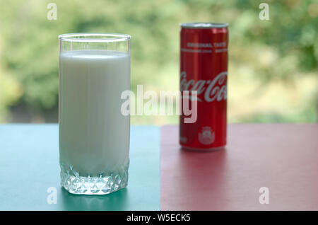 KHARKIV, UKRAINE - JULY 20, 2019: Coca cola red tin can and cup of fresh milk on colored surface with background of green garden. Healthy and unhealth Stock Photo