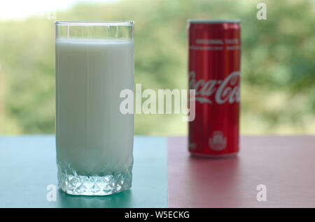 KHARKIV, UKRAINE - JULY 20, 2019: Coca cola red tin can and cup of fresh milk on colored surface with background of green garden. Healthy and unhealth Stock Photo