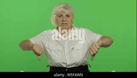 An elderly woman showing no and giving her thumb down. Old pretty grandmother in a white shirt. Place for your logo or text. Chroma key. Green screen background Stock Photo