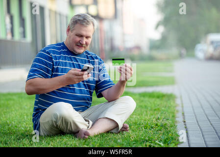 A satisfied man makes online purchases using a credit card and a smartphone, sitting on the lawn in the city Stock Photo