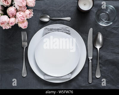 Beautiful table setting on gray linen tablecloth. Festive table setting for wedding dinner with pink spray roses and scented cabdle. Holiday dinner with white plates. Dinner, utensil. Copy space Stock Photo