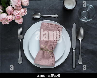 Beautiful table setting with pink spray roses and candle on gray linen tablecloth. Festive table setting for wedding dinner with pink napkin on plate. Holiday dinner with white plates Stock Photo