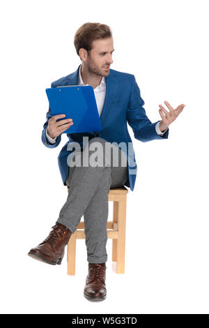 Angry businessman explaining and gesturing while frowning to the side and sitting with his legs crossed on white studio background Stock Photo