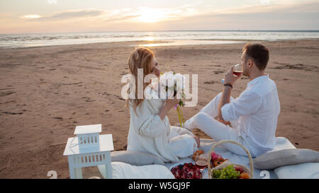 Warm sunset. A man and a pregnant woman had a picnic on the sand with a blanket, pillows, a lantern, fruit and sweet pastries. Stock Photo